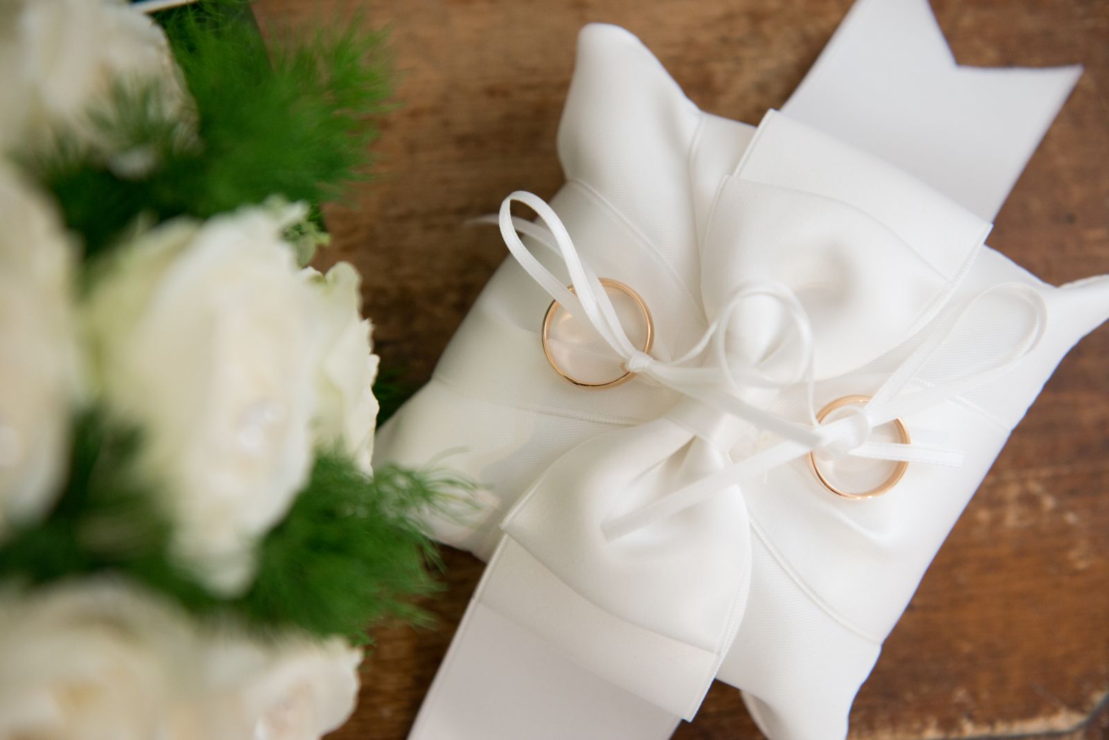 Le fedi nuziali appoggiate su un cuscino di raso accanto a un bouquet di rose bianche
