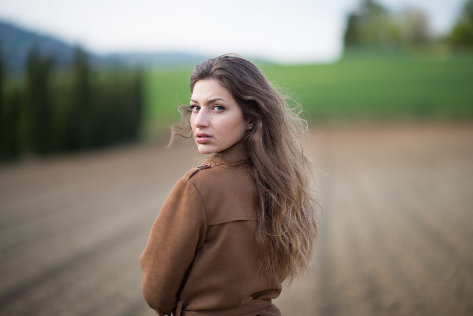 Ritratto di ragazza con cappotto cammello, sguardo verso l'obiettivo, sfondo di campagna sfocato